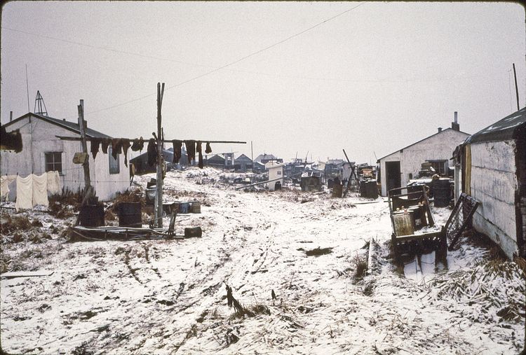 Okpowruk's homes, now gone due to erosion, located on west end of town. Later, newer homes were constructed just west of these homes but they were all relocated to the old airport site (which isn't used now).  If you were there now, where the picture was taken, you would be standing on the same sand but maybe surrounded by water. Charlie Okpowruk's home had been seen world-wide as it lay sideways at an angle. Now the building has been lost to the sea.