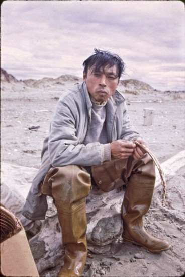 Morris “Moe” Kiyutelluk sitting on a drift log, snacking on dried caribou or dried reindeer. He is known as the “walking, talking historian for Shishmaref.”